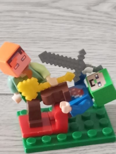 The image displays a collection of Minecraft-themed toys arranged on a wooden surface. In the foreground, two figures are depicted. One figure, with an orange head and a green torso, is in a reclined position, its legs extended and body tilted. It holds a yellow, flower-like object in its right hand and appears to be interacting with the other figure. The second figure, wearing a blue shirt and a green helmet or headpiece, is positioned beneath the first. Its head is tilted upwards, and its right hand is raised, possibly in defense or interaction. A grey, pixelated sword is visible in the upper portion of the image, positioned above the figures, suggesting a playful or combat-related scene.
The figures are constructed from plastic, characteristic of building block toys. They are placed on a green baseplate with raised studs, a common feature of construction toys. Other red and brown plastic elements are also integrated into the scene, forming part of the figures' attire or accessories, and possibly a small platform. The background consists of a light-colored, wooden surface with visible grain, suggesting an indoor setting. The lighting appears to be soft and even, with no strong shadows, indicating it might be daytime or a well-lit indoor environment. There are no visible text elements in the image. The overall impression is a staged play scene with Minecraft characters.