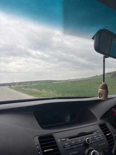 The image shows the interior view of a car, focusing on the dashboard, windshield, and rearview mirror. Through the windshield, a road is visible with a grassy field on one side and a cloudy sky above. Hanging from the rearview mirror is a wooden car freshener with the word "neon" engraved. The dashboard includes a car stereo system with buttons for AM, FM, CD, and AUX, as well as controls for volume and track selection. The stereo display shows the time as "17:36" and the radio station as "FM2 99.00". Due to the limited visual information, the location cannot be confirmed to be Spahievo, Bulgaria.