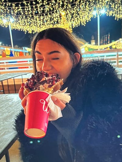 A young woman is holding a large waffle cone filled with ice cream, toppings, and chocolate sauce. She is wearing a black fuzzy coat and is in an outdoor setting with many string lights hanging overhead, suggesting it is nighttime and likely a festive event or market. The background is blurred but appears to include buildings and possibly an ice skating rink. The woman's eyes are closed as she brings the waffle cone to her mouth. A red cup with a white logo is visible in her hand, along with a white napkin. The image does not provide any specific information about the city of Chotěšov, Czechia, or its location within the city.