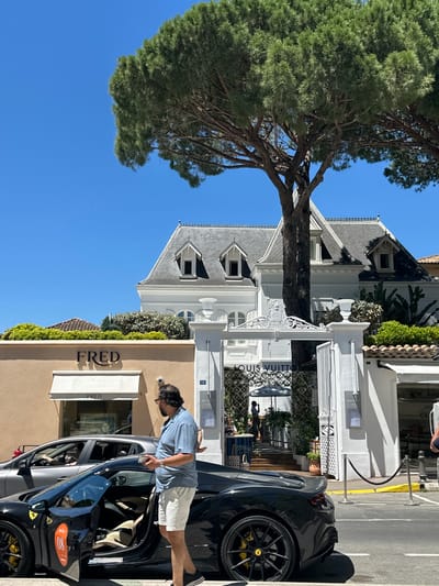 A man stands on a sidewalk in Saint-Tropez, France, holding a white cup and his phone. He is wearing a blue and white striped short-sleeved shirt and white shorts. Next to him, a black sports car with its driver's side door open is parked on the street. The car has yellow brake calipers and an orange circular decal on its side with the number "08". Behind the man is a building with a tan facade featuring the "FRED" logo and a white awning. Further back, a white building with "LOUIS VUITTON" signage and an ornate gate is visible, with lush green foliage and trees surrounding it. The sky is clear blue, indicating a sunny day.