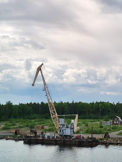 The image depicts a stationary crane on a barge floating in a body of water in Davydovskaya, Russia. Behind the barge, there is a green landscape with trees and grass. A small wooden house is visible to the right of the scene. The sky above is filled with clouds.