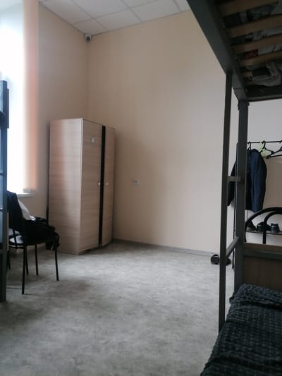 The image shows a sparsely furnished room, possibly a dormitory or shared living space, in Комсомольский, Russia. A light-colored wardrobe with two doors stands against the back wall, to the left of a wall-mounted electrical outlet. To the left of the wardrobe, a chair with some clothes draped over it is positioned near a window with vertical blinds. A security camera is visible in the upper left corner of the room, mounted on the ceiling. In the right portion of the image, a metal bunk bed frame is present, with some bedding and clothing visible on the upper bunk. A clothes rack with jackets and hangers is mounted on the wall behind the bunk bed. A pair of sandals rests on the floor near the bunk bed. The floor is covered in a gray, textured material. The walls are painted a neutral, light color. The overall impression is one of a functional, unadorned living space.