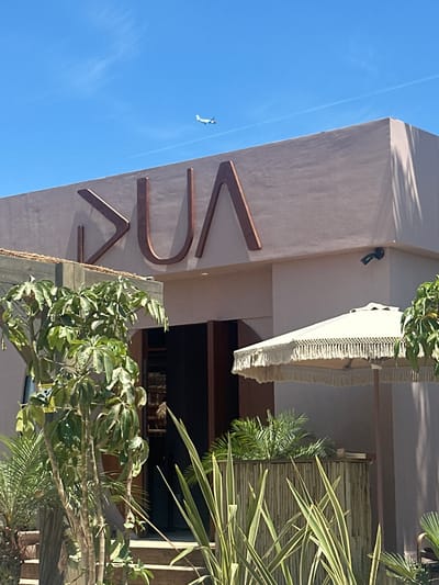 An external, eye-level, medium shot captures the facade of a building with prominent copper lettering spelling out "DUA" against a pale pink wall under a clear blue sky. An airplane is visible flying high in the sky, leaving a faint contrail. To the right of the building entrance, a white patio umbrella with decorative fringe is positioned over a bar constructed from bamboo. Lush green plants, including a palm tree and a leafy plant with mottled leaves, are in the foreground. Wooden stairs lead up to the entrance of the building. A security camera is mounted on the exterior wall to the right of the entrance. The scene appears to be in Guadalmar-San Julián, Spain.