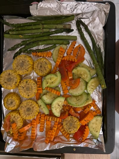 The image shows a baking sheet lined with aluminum foil, filled with a colorful assortment of prepared vegetables. The vegetables appear to be seasoned and ready for cooking.

Prominently featured are several stalks of asparagus, arranged vertically on the left side of the sheet. Scattered amongst them are slices of zucchini, cut into half-moons, and bright orange carrot sticks, many with a crinkle-cut edge. There are also rounds of corn on the cob, some with visible kernels. Pieces of red bell pepper can be seen interspersed with the other vegetables.

The vegetables are arranged loosely across the baking sheet, suggesting they are being prepared for roasting or baking. The lighting is bright, indicating it's likely daytime, and the scene is likely in a kitchen or other indoor food preparation area. There are no visible people, text, or specific location cues to suggest a time of day, weather, or emotion, beyond the preparation of a meal. The overall impression is one of healthy, ready-to-cook ingredients.