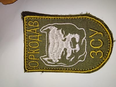 The image displays a close-up view of an embroidered patch. The patch is rectangular with rounded corners and a dark olive green background fabric. It features a gold embroidered border and lettering. In the center, there is a white embroidered outline of an animal, possibly a horse or deer, with internal stitching. To the left of the central image, in gold lettering, are the Cyrillic letters "ОРКОЛАБ". To the right of the central image, also in gold lettering, are the Cyrillic letters "ЗСУ". The patch appears to be a military or organizational insignia. The context of Okhtyrka, Ukraine, is not visually present in the image itself.
