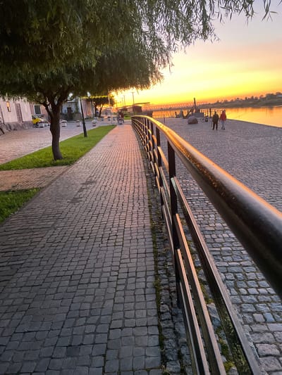This image depicts a cobblestone promenade alongside a river at sunset in Sovetsk, Russia. A dark metal railing runs along the right side of the promenade, reflecting the warm hues of the sky. On the left, a tree with drooping branches overhangs a grassy area and a cobblestone street, where a yellow car is parked. Further down the promenade, two individuals are walking away from the camera, their figures silhouetted against the vibrant sunset. The river stretches across the background, with a bridge visible in the distance and a tree-lined shore on the opposite bank. The overall scene conveys a peaceful, late-evening atmosphere.
