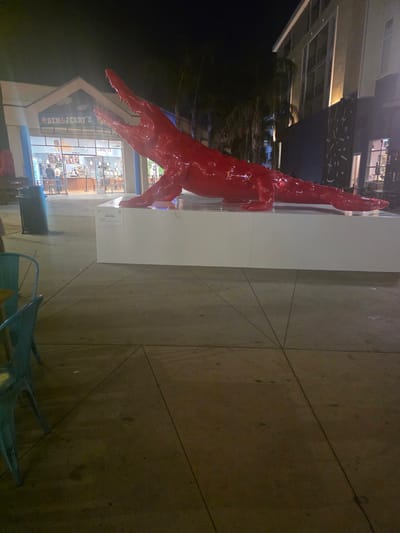 A large, bright red, glossy sculpture of a crocodile is displayed outdoors on a white rectangular pedestal at night. The crocodile sculpture is positioned horizontally with its mouth open, revealing sharp teeth. It is situated on a paved walkway, with a building housing a Ben & Jerry's ice cream shop visible in the background. The building has large glass windows and doors, with some patrons visible inside. To the right of the sculpture, a modern building with illuminated windows and balconies is present, featuring a dark facade with abstract white patterns. In the foreground, to the left, are outdoor seating areas with teal-colored chairs and tables. The lighting suggests it is evening, with artificial lights illuminating the scene. The location is identified as being in Burbank, United States of America.
