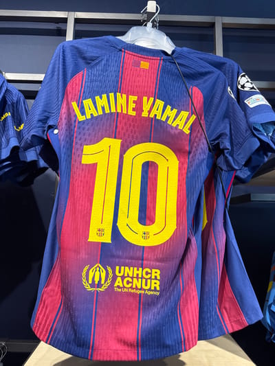 The image shows the back of a blue and red striped FC Barcelona jersey with "LAMINE YAMAL" printed in yellow lettering above the number "10". Below the number, the UNHCR logo is visible. The jersey is hanging on a display rack in a retail setting, likely a store. Another jersey with similar design is partially visible to the left. The background consists of dark shelving and a dark wall. The scene is well-lit. The location of La Roca del Vallès, Spain is implied by the context of an FC Barcelona merchandise display.