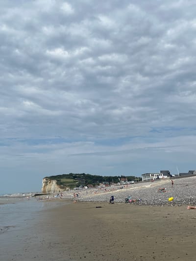 The image depicts a view of a beach and cliffside with a town or resort area. The scene is oriented vertically, with the sky occupying the left portion of the image and the beach, town, and cliffs on the right.

The sky is filled with a dramatic, overcast cloud cover, with some lighter patches suggesting diffused sunlight. The sea is not prominently visible, but the wet sand of the beach indicates that the tide is either out or receding. The beach itself is a mixture of sand and pebbles, with scattered people enjoying the area.

Along the edge of the beach, there is a rocky embankment or seawall, behind which are buildings, some appearing to be modern resort accommodations, and older structures. Further inland, a green, treed cliff rises, suggesting the geographical context. There are many people visible along the promenade or beach, some sunbathing on chairs, others walking or standing. A bright yellow object, possibly a beach ball or toy, is visible on the pebble area.

The overall impression is that of a moderately busy beach on a cloudy day. The location cues, such as the architecture and the cliff formation, along with the given context, point to a European coastal town. There is no visible text in the image.
