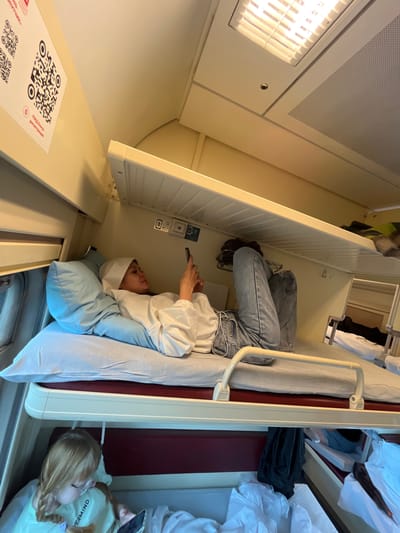 A medium shot captures a woman lying on a top bunk in a train compartment, illuminated by overhead fluorescent lights. She is wearing a white hoodie and blue jeans, with a white headscarf, and is engrossed in her smartphone. A large blue pillow supports her head. Below her, on the lower bunk, a young girl with blonde hair in braids, wearing glasses and a light green t-shirt, is also looking at a smartphone. The train compartment features cream-colored walls, beige bunks with red accents, and metal safety rails. QR codes and informational text are visible on the wall to the left. The overall environment suggests a travel setting, likely on a train within Russia, given the Cyrillic text on the QR codes. The image does not provide specific details to confirm the location within the city of Vyatskiye Polyany, Russia.