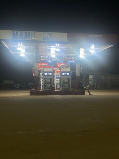 Here is a description of the image:

The image shows a Mamu Oil gas station located in Kofan Doka Zaria, Unguwar Fatika, Nigeria at night. It depicts three gas pumps numbered Pump 1, Pump 2, and Pump 6. Several vehicles are visible parked near the pumps. A person is standing between the pumps and the building.