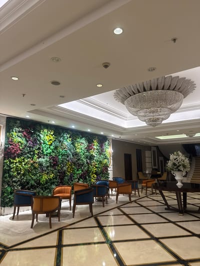 The image shows an interior space in what appears to be a hotel or large building in Bağcılar, Turkey. A large, ornate chandelier hangs from the ceiling. Along one wall is a vertical garden or wall of greenery. There are a row of chairs upholstered in alternating blue and orange fabric, arranged near the green wall. The floor is tiled in a pattern of light colored stone with dark accents forming a grid. The overall impression is of a modern, well-lit lobby or waiting area.