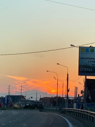 A low-angle, eye-level shot captures a street scene at sunset in an urban environment. The sky is a gradient of pale yellow at the top transitioning to vibrant orange and red hues near the horizon, with wispy clouds illuminated by the setting sun. A power line stretches across the upper portion of the frame.
In the foreground, a paved road curves to the left, with white dashed lines marking its lanes. A metal guardrail lines the right side of the road.
The middle ground features several streetlights with curved arms, some silhouetted against the sky, along with utility poles and wires. Buildings with dark roofs are visible, and a tall tower structure with multiple levels stands on the left. A billboard with blue, green, and yellow logos and Cyrillic text is positioned on the right side of the frame. Partially obscured text on the billboard includes "КВАРТИРА" (Apartment) and a phone number "03711".
In the background, more buildings and a distant silhouette of a church or similar structure can be seen. The overall atmosphere is one of an evening approaching in a moderately developed urban area.