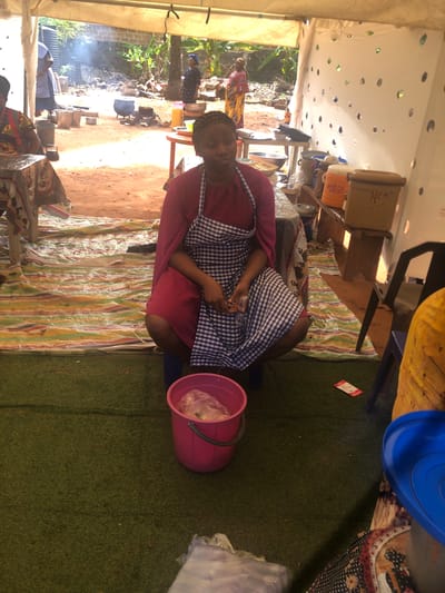 In Nteje, Nigeria, a woman wearing a red top and a blue and white checkered apron is seated under a tent. She is holding something in her hands, and a pink bucket is beside her. Outside the tent, there's a cooking area with pots on open fires. The tent's roof has numerous small holes, and the floor is covered with a striped rug and artificial turf.