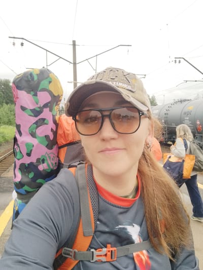 A selfie is taken by a woman on a train platform in Orekhovo, Russia. She is wearing a tan baseball cap and dark-rimmed sunglasses. She has long, reddish-brown hair and is looking directly at the camera. She is wearing a grey long-sleeved shirt with an orange collar and a backpack with orange straps. To her left, a large, colorful bag with a camouflage pattern of pink, green, yellow, and black is leaning against her. Behind her, a train car with "wpt" printed on it is visible. In the background, another woman with blonde hair is walking away from the camera, carrying a green backpack and two orange bags. The platform is marked with yellow lines, and train tracks are visible to the left. The sky is overcast.