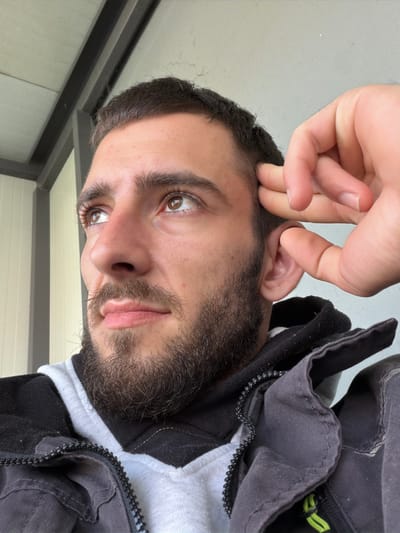 A close-up, slightly angled portrait shows a bearded man indoors. His left hand is raised near his left ear. He has brown hair and eyes. The background includes sections of a wall and part of what appears to be clothing. There is no information in the image that indicates the location is in Yoakim Gruevo, Bulgaria.