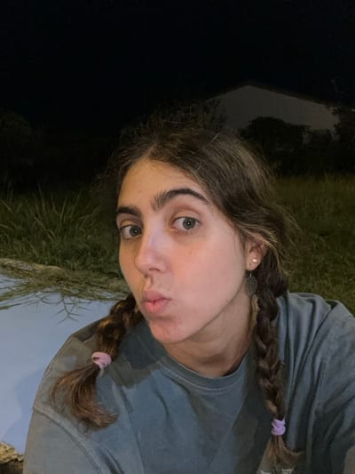 A young adult female is depicted in a close-up, waist-up shot at night in Thap Sakae, Thailand. She has long, wavy, medium-brown hair styled into two braids, each secured with a light purple elastic band. Her light-colored eyes, appearing blue or green, gaze directly forward, and her lips are pursed in a pout. She wears a plain, short-sleeved, light grey or faded olive-green t-shirt. Her visible right ear features a small stud earring in the upper lobe and a larger, dark, ornate dangle earring in the lower lobe. The background is dimly illuminated, revealing patches of tall green grass in the lower left and a light-colored, possibly concrete, surface. In the upper right, the dark silhouette of a structure with a sloped roofline is visible against the dark night sky, partially obscured by dark foliage.