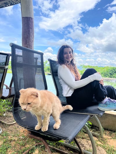 A young woman with curly brown hair is seated on a black outdoor chair. She is wearing a white long-sleeved shirt and black pants. Her left arm is resting on her knee, and she is looking at the camera with a smile. In front of her, on another black outdoor chair, is a light orange cat. The cat is standing and looking down. The chairs are situated outdoors next to a body of water, possibly a pool or a lake, with lush green vegetation and distant mountains visible under a partly cloudy sky. The location is specified as Sơn Điền, Vietnam. The scene appears to be a relaxed, outdoor setting.