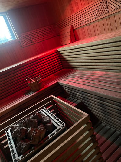 This image displays the interior of a sauna room, identified as being located within Fraccionamiento y Club de Golf los Encinos, Mexico. The room's walls and multi-tiered benches are constructed from light-toned horizontal wooden planks. Triangular patterns formed by angled planks are visible on the upper wall panels. A dominant red light emanates from an unseen source, likely LED strips, positioned along the lower wall sections and underneath benches, casting a crimson glow throughout the space, particularly on the left side. A rectangular window is present in the upper left, providing ambient external light. In the immediate foreground, a sauna heater is enclosed within a slatted wooden frame, containing multiple dark, irregular rocks resting on a light-colored metal grate. On a higher bench to the left, a small wooden bucket holds a wooden ladle. No individuals, activities, or interactions are depicted in the static scene.