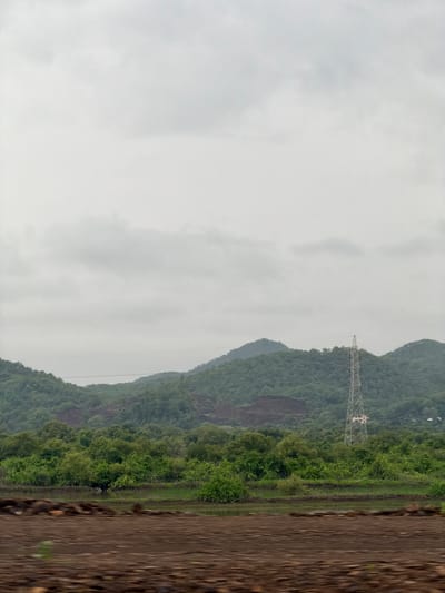 The image depicts a landscape with rolling green hills under a cloudy sky. In the foreground, there is a dirt field with some blurred brown objects, possibly rocks or debris. Beyond the field, there is a body of water, appearing as a marshy area with dense green vegetation lining its banks. In the middle ground, a dense forest with various shades of green foliage stretches across the landscape. A tall, metallic power transmission tower stands prominently to the right of the frame, silhouetted against the green hills. The background features more undulating hills covered in lush vegetation. The overall scene suggests an outdoor environment, possibly in a rural or semi-rural area. The location is identified as Vasai-Virar, India.