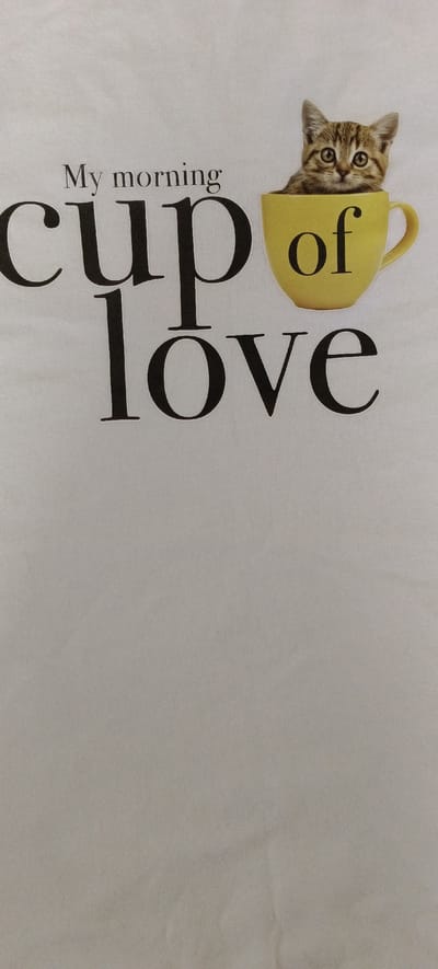 A close-up photograph displays a white t-shirt with a graphic design. The graphic features the text "My morning cup of love" in a stylized font. A small, tabby kitten is pictured peeking out of a yellow coffee cup. The text is arranged in three lines, with "My morning" at the top, "cup" in large letters below, and "love" in even larger letters at the bottom. The word "of" is printed in smaller letters within the yellow cup. The t-shirt is likely being worn by someone in a domestic setting or is a standalone garment. No specific location in Падлабенскі сельскі Савет, Belarus is identifiable from the image.