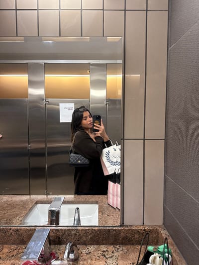 An image taken in a public restroom in Lawrence Township, United States, depicts a young adult female captured in a mirror selfie. She has dark hair and wears a dark, long-sleeved top. A black quilted shoulder bag with a gold chain strap hangs from her left shoulder. She holds a dark-colored smartphone, featuring a gold-toned case with the word "CHANCE," up to take the photo. Her right arm carries several shopping bags, including a pink Victoria's Secret bag and a white bag with a blue wreath design. The restroom environment features a light brown speckled granite countertop with an integrated white sink basin and a chrome automatic faucet. The mirror reflects three stainless steel toilet stall doors, one displaying an inverted "Temporary Closed" sign, and warm yellow lighting positioned above them. The wall above the mirror consists of light beige square tiles, while the wall to the right of the mirror has a textured grey surface. On the countertop to the far right, a black basket holds various green and white items.