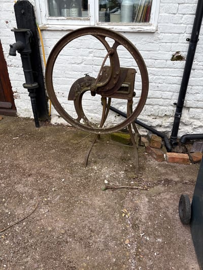 The image, oriented horizontally, features a vintage chaff cutter positioned against a white brick wall. The chaff cutter is made of rusted metal and has a large, circular wheel as its primary feature. The mechanism of the cutter includes blades and a feed trough.
The wall is part of a building exterior, marked with signs of age and weathering. A window frame, painted white, is visible to the left of the cutter. Above the cutter, black pipes run along the wall.
To the right of the wall, there is a paved area with a rough, textured surface, suggestive of a yard or outdoor space. A wheel, likely from a bin, can be seen in the top right corner.
A yellow hose is present below the chaff cutter. Also below the chaff cutter is a water pump mounted on a black wooden base.
The scene is likely located in Longden, United Kingdom. No people are visible in the image.