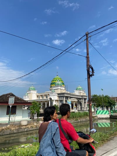 The image shows a scene in Kecamatan Tulangan, Indonesia (latitude: -7.475, longitude: 112.634). A man and a woman are sitting on a red moped. The man is wearing a red shirt and black shorts. The woman is wearing a blue jacket over a green shirt. They are both looking to the right. Behind them is a mosque with a green and yellow dome. The mosque is surrounded by a canal. The sky is blue with some clouds. There are power lines running across the sky. There are lush green plants around the canal and a small green sign in the foreground.