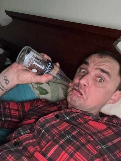A man in a red and black plaid shirt is lying in bed, holding a glass bottle to his nose. The bottle has a label that reads "1 БАЙКАЛ". The man's eyes are wide open and his mouth is contorted. The background shows a wooden headboard and a pillow. There is also a tattoo of an infinity symbol on the man's left wrist. The scene is consistent with a bedroom in a residential building in Lesnoi Gorodok, Russia.