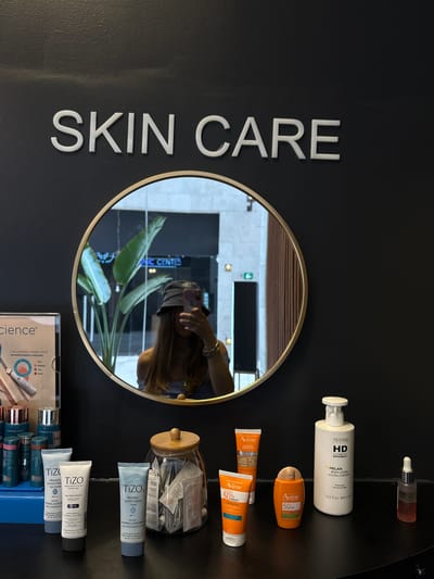A vertically-oriented photograph shows a display of skincare products, with a reflective, circular mirror in the center. To the left of the mirror, large white lettering spells out "SKIN CARE" vertically. Various skincare items are arranged to the right of the mirror including tubes and bottles of sunscreen and lotions. Brand names such as "Avène" and "TiZO" are visible on the products. There is a jar filled with small packets, possibly samples, and a color cosmetics display tray at the bottom right. The mirror reflects the photographer holding a cell phone, a potted plant, and the exterior of a business identified by a sign that includes the words "IC CENTER", which places the scene in Ciudad López Mateos, Mexico.