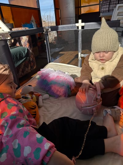 The image shows two children lying on what appears to be a bed or padded surface, with various objects around them. One child is wearing a brown outfit with a pointed knit hat and is holding a small pink purse shaped like a basketball. The other child is wearing pink and teal pajamas with a heart pattern. Near them are a colorful tutu, a stuffed animal resembling a tiger, and other unidentifiable objects. The scene is situated indoors, possibly within a screened enclosure. The location is identified as Кипчак, Russia, although this detail cannot be visually confirmed within the image.