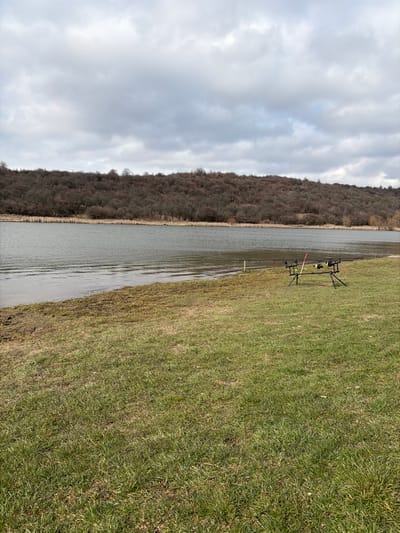 The image presents an outdoor scene depicting a body of water and a forested hill, situated in Cherna Gora, Bulgaria. The foreground comprises a stretch of green grass, interspersed with areas of drier, lighter vegetation, terminating at a muddy, sparsely vegetated bank bordering the water. A black, three-legged fishing rod pod, holding three dark fishing rods equipped with electronic bite alarms (one with a yellow indicator, one with a red), is positioned on the grass to the right of the frame's center, facing the water. Two slender, yellow-tipped marker poles are partially visible near the water's edge; one to the left of the rod pod and another further to the right. The midground features a calm, greenish-grey body of water with minor surface ripples. The opposing bank is lined with tall, dried reeds. The background consists of a gentle hillside densely covered with leafless deciduous trees, exhibiting a reddish-brown coloration. The sky is uniformly overcast with grey cloud cover. No individuals are present.