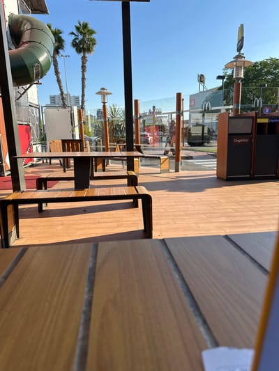 The image shows an outdoor seating area at a McDonald's restaurant in Sedaví, Spain. The scene is captured during daylight with a clear blue sky. In the foreground, wooden picnic tables and benches are visible, with the floor made of wooden planks. Behind the seating area, a play structure with a green tube slide is prominent, along with palm trees and modern buildings in the background. A recycling station with labels for "Orgánico" (organic) and "Plástico/Líquidos" (plastic/liquids) is also present. The environment appears clean and well-maintained, characteristic of a fast-food establishment.