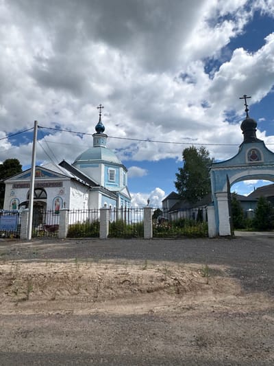 The image shows an Orthodox church with a blue dome and cross, located in the village of Глебовское, Russia. The church is white with blue trim and features several religious icons painted on its exterior walls. A fence with decorative pillars encloses the church grounds, where a small garden with flowers is visible. To the right of the church, a blue archway with a religious icon on top serves as an entrance. The church is situated on a gravel road, with a dirt embankment in the foreground. The sky is partly cloudy with a mix of blue sky and white clouds.