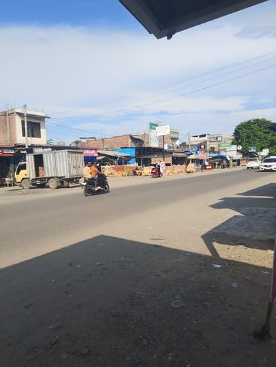 A medium shot captures a street scene in Gampongpaya, Indonesia, under a bright, partly cloudy sky, suggesting it's midday. The foreground is dominated by a dusty, unpaved shoulder of a road casting a long shadow from an unseen overhang. The road itself is paved and shows signs of wear.
On the road, a yellow truck with a large, silver cargo container is parked. Inside the container, boxes are visible. A black motorcycle carrying two men in casual attire, one wearing an orange shirt and the other a lighter orange shirt, rides past the truck, heading away from the camera. The rider of the motorcycle is wearing a helmet.
The background features a row of buildings that appear to be shops and food stalls. Many have colorful awnings and signs. Some of the visible text on the signs includes "NYALARAN MERA YANG MELEGEND," "JIMSUM msum Premium 2.000," "ES TELER CREAM DURIANIED," "ES TELP," and "OPPO." This suggests a commercial area with food vendors and perhaps electronic shops.
Further down the street, more buildings and stalls are visible, some with red and blue awnings. Two white cars are parked on the side of the road near a lush green tree. The overall impression is of a bustling, everyday street scene in a tropical or subtropical region, characterized by a mix of commercial activity and transportation.