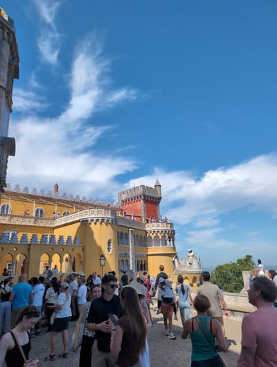 The image shows the Pena Palace in Sintra, Portugal. A group of tourists stands near the palace, some interacting with each other and their phones. The architecture of the palace is prominently displayed, with its yellow and red facade, ornate details, and towers. The sky is blue with some clouds.