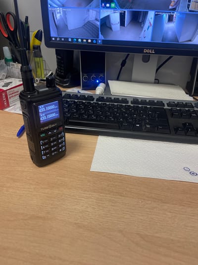 A walkie-talkie with a digital display rests on a wooden desk in front of a computer monitor displaying multiple security camera feeds. The walkie-talkie shows two frequencies: 433.10000 MHz and 433.15000 MHz. A keyboard and several rolls of paper towels are also visible on the desk. In the background, a black phone sits in a charger. The scene appears to be an office or control room. No people are visible in the image. The image appears to have been taken in Obninsk, Russia, based on the presence of a Klever brand product, which is a Russian stationery brand.