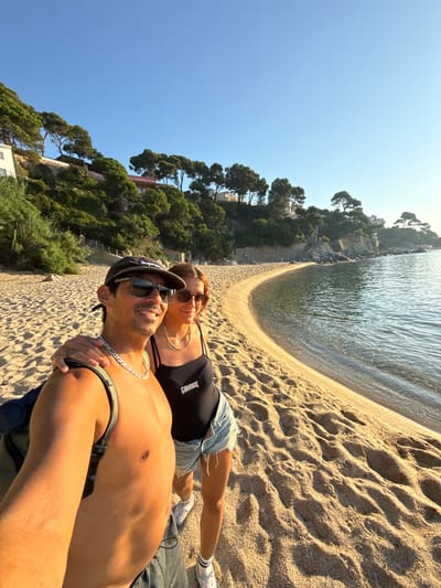 A man and a woman are taking a selfie on a sandy beach in Castell d'Aro, Platja d'Aro i s'Agaró, Spain. The man, shirtless and wearing a baseball cap and sunglasses, has his arm around the woman. She is wearing a black swimsuit and denim shorts. The beach is golden sand with footprints, and the clear water of the Mediterranean Sea gently laps at the shore. In the background, a lush, green hillside with scattered trees and buildings rises steeply, characteristic of the Costa Brava coastline. The sky is a clear, bright blue, indicating a sunny day. The overall scene suggests a relaxed vacation atmosphere.