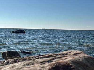The image depicts a tranquil seascape under a clear blue sky. In the foreground, a large, textured rock dominates the lower portion of the frame, with another smaller rock partially submerged in the water to its left. The sea is calm with gentle ripples, reflecting the bright sky. In the middle ground, a larger rock emerges from the water, with a solitary white bird perched on its surface. Further in the distance, on the horizon, a ship is faintly visible, appearing as a small silhouette against the bright sky. The overall atmosphere is serene and natural, suggesting a peaceful outdoor setting. The location is Rannamõisa, Estonia.