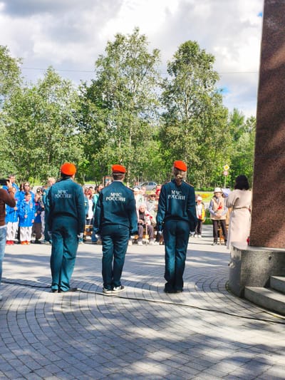 In Apatity, Russia, a group of people are gathered outdoors, possibly for a commemorative event. Three individuals in the foreground wear matching dark blue uniforms with orange berets. Their uniforms bear the Cyrillic text "МЧС РОССИИ" on the back, indicating they are part of the Russian Ministry of Emergency Situations. They stand facing away from the camera, towards a crowd of onlookers, suggesting they are on duty or participating in a formal capacity.

The crowd consists of people of various ages, some dressed in casual attire, others in more formal wear, and some children. Many are looking towards the individuals in uniform or towards something beyond the frame, hinting at a ceremony or a moment of reflection. Some individuals are holding small red flowers, commonly used in memorial services.

The setting appears to be a paved public space, possibly a town square or park, with a large, dark monument to the right of the frame. Trees with green foliage form a backdrop, and the sky is partly cloudy with scattered sunlight, indicating it's likely daytime, perhaps late morning or afternoon. The overall mood seems respectful and solemn.