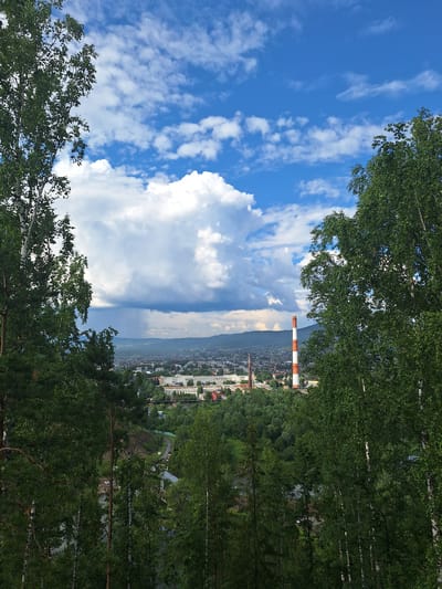 The image shows a panoramic view of the city of Sim, Russia, framed by lush green trees in the foreground. A tall, red and white striped industrial chimney stands prominently in the middle ground, amidst a cluster of buildings that make up the city. The sky above is a vibrant blue, dotted with fluffy white clouds, some of which appear to be gathering into a storm system in the distance, with visible rain streaks. The overall scene depicts a blend of natural landscape and industrial presence within the urban environment.