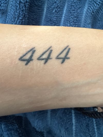 A close-up photograph shows a person's arm, presumably a forearm, with a tattoo of the number "444" vertically aligned on the skin. The tattoo is done in black ink. The arm is resting on a textured blue fabric. A dark bracelet is partially visible on the wrist. The location of the photograph is not evident from the image and cannot be determined to be in Glojene, Bulgaria, or any other specific location.
