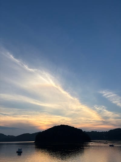 This image captures a sunset over a body of water, likely Lake Rożnowskie, near Gródek nad Dunajcem, Poland. The sky is a gradient of deep blue at the top, transitioning to lighter blues, with streaks of white and yellow clouds catching the setting sun's light. A prominent, dark, silhouetted island sits in the middle ground of the water. The water reflects the colors of the sky and the silhouette of the island. In the foreground, a small, white floating structure with a roof is visible on the water. Further in the distance, on the far side of the water, the dark silhouettes of hills and treelines line the horizon, with some scattered lights indicating human habitation. Several small boats, some appearing to be kayaks or canoes, are also visible scattered across the water, with people in them. The overall scene is serene and depicts the natural beauty of the landscape at dusk.
