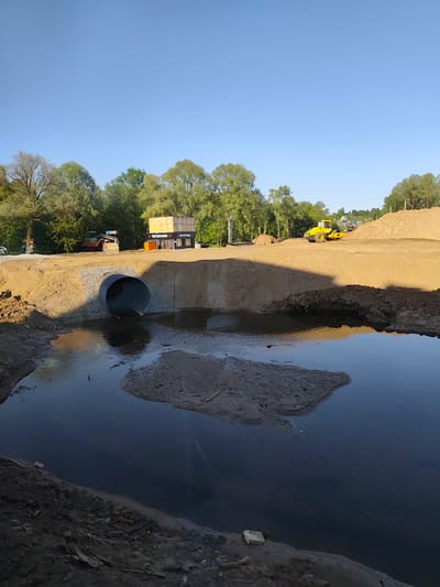 The image depicts a construction site in Oktyabr'skii, Russia. A large concrete culvert, presumably for drainage, is visible in the center of the frame, with one end submerged in a body of water, reflecting the sky. The culvert is embedded in a mound of tan-colored sand and dirt. Various pieces of construction equipment are present, including a yellow road roller on top of the mound and a dump truck parked on the elevated surface behind the mound. A structure with Cyrillic writing is also seen in the background. Trees line the horizon under a clear, light blue sky. The foreground is dominated by the body of water, showing muddy banks and a sandbar.