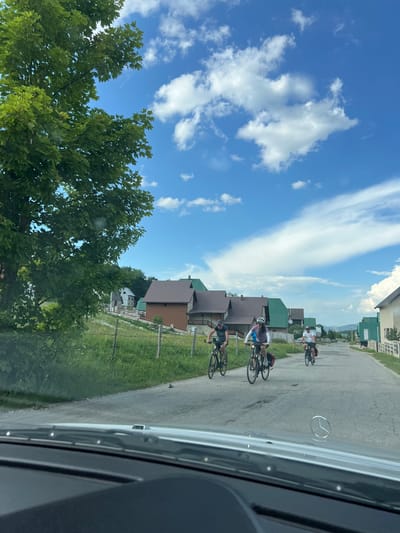 The image captures a rural scene in Motički Gaj, Montenegro, with a focus on a paved road lined by wooden houses with green and brown roofs. In the foreground, the hood and windshield wipers of a car are visible, suggesting the photo was taken from inside a vehicle. A Mercedes-Benz emblem is visible on the hood. Three cyclists are seen riding on the road, heading away from the camera. They are on separate bicycles, with the middle cyclist wearing a helmet and a purple top, and the cyclist to their left wearing a dark cap and shirt. The environment is lush with green grass and trees, under a bright blue sky with scattered white clouds. The overall atmosphere is one of a leisurely outdoor activity in a scenic, possibly mountainous, location.