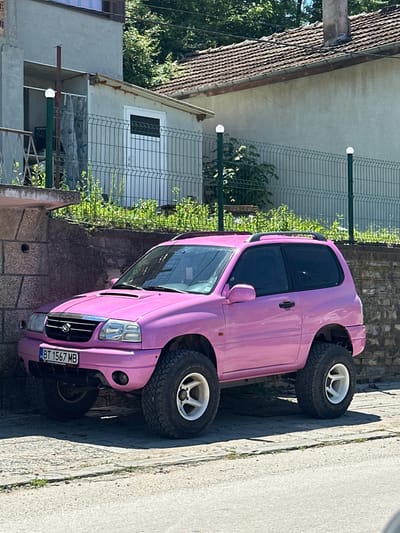 A bright pink, lifted Suzuki Vitara SUV is parked on a cobblestone surface in front of a stone wall. The vehicle has large, knobby tires and white rims. A Bulgarian license plate, "BT 1567 MB," is visible on the front. Behind the car, a metal fence with solar-powered lights runs along a retaining wall covered in greenery. Further back, buildings with tiled roofs and a chimney are visible. The scene is set on a sunny day, with shadows cast on the ground. The location is identified as being in the city of Debelets, Bulgaria.