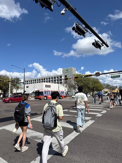 This image depicts a sunny daytime scene at a busy urban intersection in Gainesville, Florida, United States. The location is the intersection of SW 13th Street, as indicated by a street sign visible on an overhead traffic light gantry.

The foreground shows three young adult pedestrians crossing the street on a white-striped crosswalk. From left to right: a female with light brown hair, wearing a black backpack, a gray and blue top, black shorts, and white sneakers, holding a white bottle; a male with dark hair, wearing a gray Under Armour backpack, a light green shirt, light gray pants, and white sneakers; and another male with dark hair, wearing a white graphic t-shirt, light blue pants, and white sneakers, slightly behind the second pedestrian. Further to the right, a group of approximately 5-7 additional young adults, some carrying backpacks, are also crossing, with one individual riding a scooter.

In the mid-ground, a red sedan is partially visible on the left lane of the street. A large red, blue, and white bus, prominently featuring "GATOR" branding and text such as "MELT AWAY LAW" and "ONLY OFFICIAL UNIVERSITY OF FLORIDA," is situated in the middle of the intersection, suggesting a connection to the University of Florida. In the background, a multi-story white building with a green "Holiday Inn" logo is clearly visible, identifying it as the Holiday Inn Gainesville-University Center. Another building with "BURRITOS" text is partially obscured behind the bus.

The sky is bright blue with scattered white cumulus clouds. Overhead traffic light gantries, painted black, support multiple traffic signals, a white security camera, and street signs. Several traffic lights are illuminated green. Palm trees and other bare or leafy trees line the streets and sidewalks. The overall environment is an active commercial and pedestrian area.