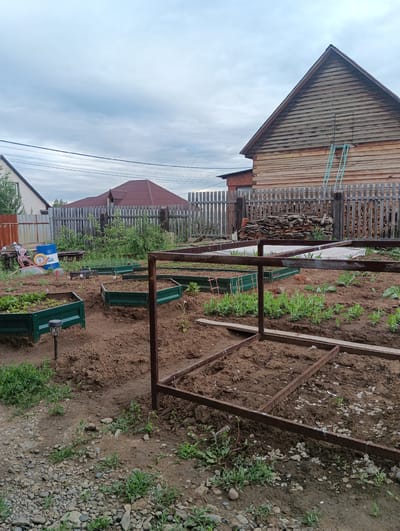The image depicts an outdoor scene in Ust-Kuda, Russia, likely a residential property with a garden. In the foreground, a partially constructed raised garden bed made of rusted metal frames is visible, filled with dark soil and some sparse green plants. Several completed hexagonal raised garden beds, painted dark green, are also present, some containing small plants. The ground is a mix of soil and gravel, with scattered green vegetation.

In the background, a weathered wooden fence separates the garden area from what appears to be a yard. Behind the fence, a pile of split logs and a stack of what looks like broken bricks or tiles are stored. Further back, a large, wooden two-story building with a dark roof and horizontal siding dominates the right side of the frame. A light blue ladder is leaning against the side of this building. To the left, a smaller building with a reddish-brown roof is visible, and beyond that, another house with light-colored siding and a dark roof. Power lines are strung across the sky. The sky is overcast with gray clouds. There are no people or animals in the image.