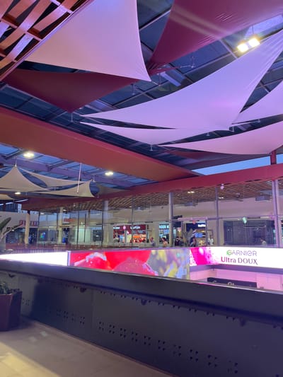 This image shows the interior of a multi-level shopping mall, likely in Mechouar Kasbah, Morocco. The view is angled upwards.

The ceiling is decorated with triangular fabric shades in white and maroon. A red and brown wooden latticework structure is visible alongside.

To the right, the mall's architecture is visible, with glass-fronted shops and walkways. There are several advertisements, including a large vertical sign for "GARNIER Ultra DOUX."  Below the Garnier advertisement there is an ad with the words "GARNIER A UR". There are also signs for "BURGER KIN" and "AVENUE". People can be seen in some of the shops and walkways.

A dark grey structural element, possibly a beam or part of the building's framework, appears in the lower right corner, partially obscuring the view.