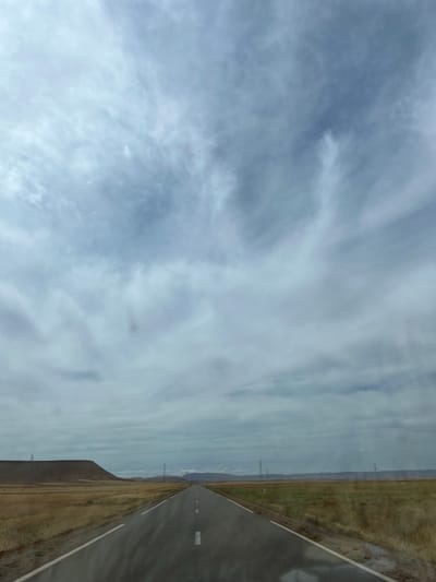 An image captures an open road extending into the distance under a sky with cloud cover. The road is flanked by land. Power lines appear along the landscape. The location is suggestive of a remote region of Sidi Chiker, Morocco.